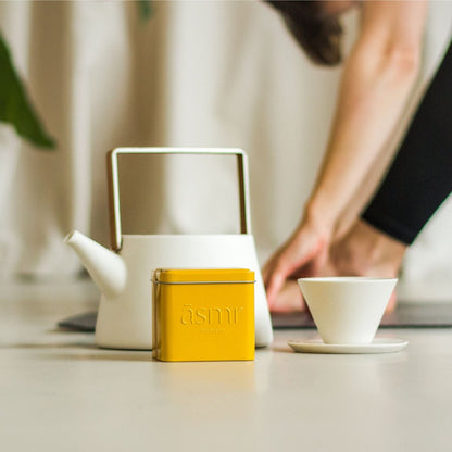 Ashwagandha Kurkuma Tee für Energie von āsmi Ayurveda mit weißer Teekanne und Tasse auf hellem Tisch