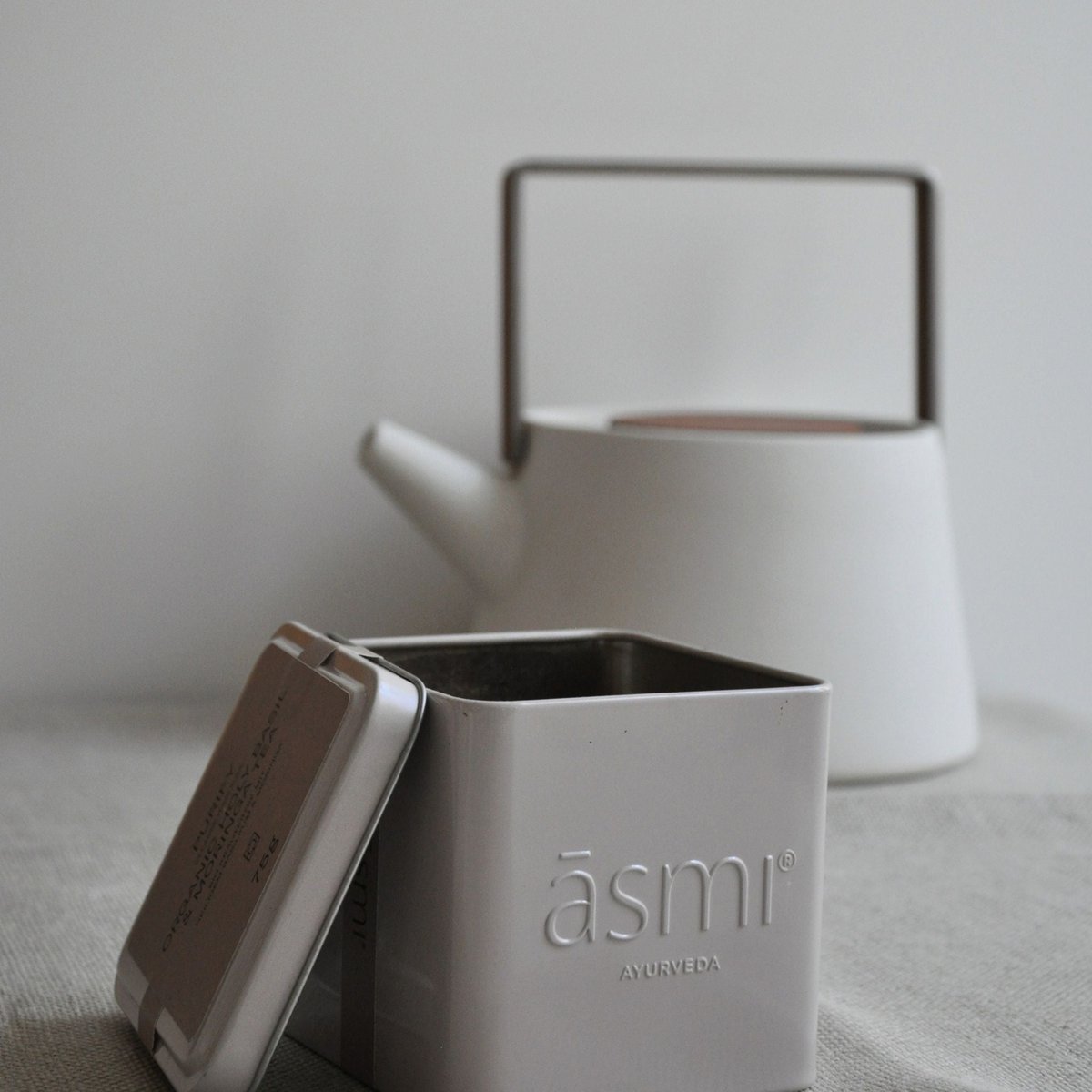 āsmi Ayurveda Blechdose mit heiliger basilikum tee bio vor weißer Teekanne auf grauem Leinentuch