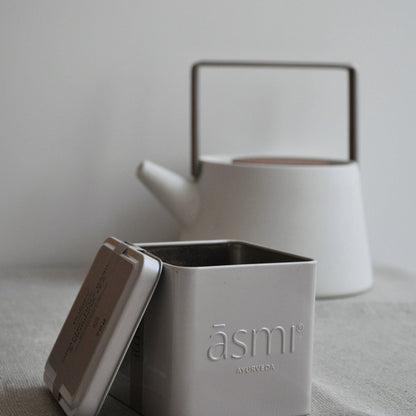 āsmi Ayurveda Blechdose mit heiliger basilikum tee bio vor weißer Teekanne auf grauem Leinentuch