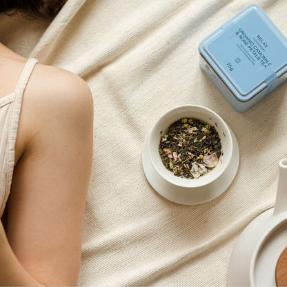 Kamillen Tee mit Rosenblütenblätter von āsmi Ayurveda in weißer Tasse neben blauer Dose und entspannter Person