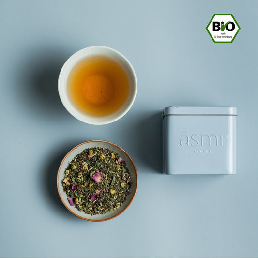 āsmi Ayurveda Kamillen Tee mit Rosenblütenblätter in weißer Dose neben aufgebrühtem Tee und losem Tee im Schälchen